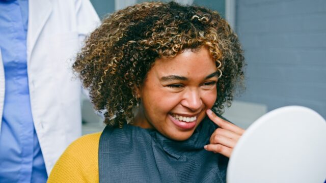 A woman with curly hair and highlights smiles broadly while touching her cheek, wearing a dark dental bib over a yellow top, seated indoors, possibly in a dental clinic.