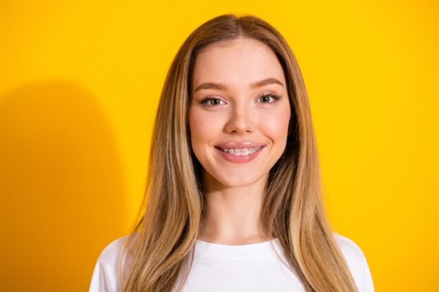 A young woman with straight, long blonde hair smiles at the camera. She wears a white shirt and stands in front of a bright yellow background. The lighting is even and her expression is friendly.